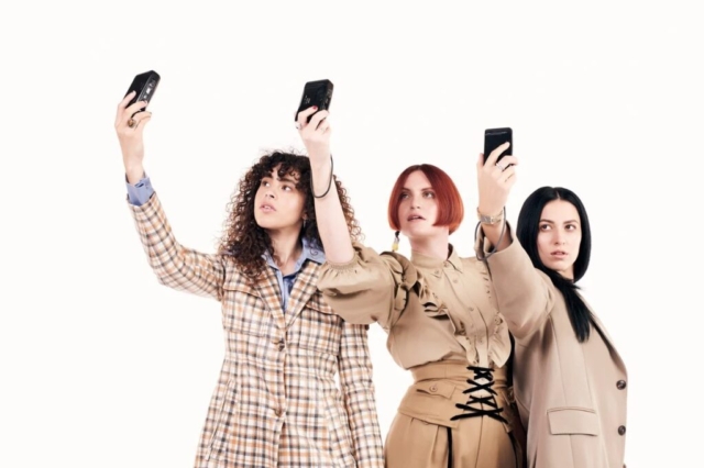 MUNA standing in a row and each holding up recorders and looking at them like they are taking a selfie.